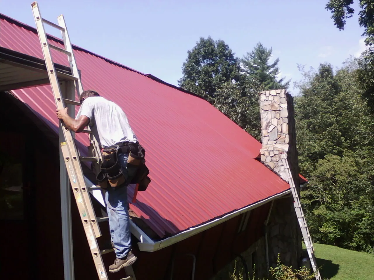 Roofer on a ladder beside a red metal roof and stone chimney — Soffit & Fascia Repair by Roofing Brothers in Etna